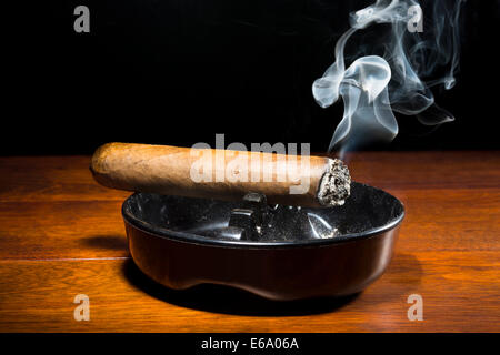 Un sigaro di masterizzazione in un classico posacenere nero fumo di streaming in un buio, moody impostazione. Foto Stock