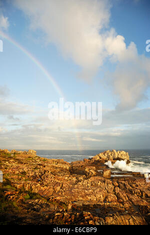 Rainbow e di onde che si infrangono sulla spiaggia rocciosa al tramonto, Hermanus, Sud Africa Foto Stock