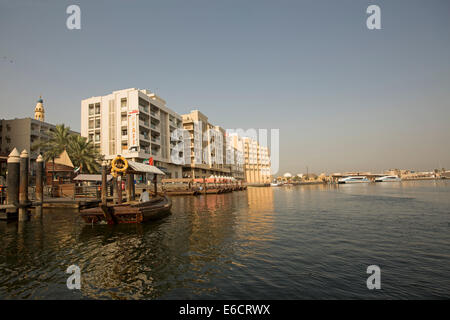 Piccola imbarcazione, abra, barca di legno tradizionale / traghetto sul Dubai Creek con elevato innalzamento edifici della città accanto a larghe vie navigabili di infilzare nel cielo blu Foto Stock