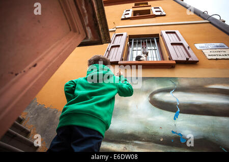 Ragazzo cerca di chiamare un gatto che è posto a sedere su una finestra nella città italiana di Dozza, Emilia Romagna, Italia, noto per pareti artistico murale Foto Stock