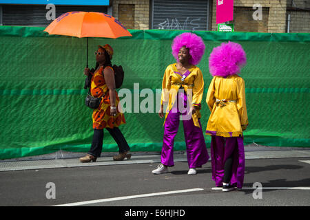 Londra, Regno Unito. 24 Ago, 2014. Due signore in viola afro parrucche e coloratissimi costumi pausa prima di prendere parte alla sfilata di Carnevale come una donna passa da con un ombrello arancione Credito: a Vista/fotografica Alamy Live News Foto Stock