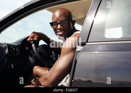 Ritratto di felice giovane ragazzo in sua automobile guardando la telecamera sorridendo. African maschio modello indossando occhiali da sole. Uomo muscolare in auto. Foto Stock