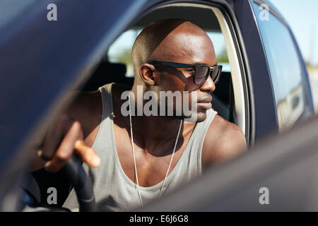 African modello maschile in sua automobile. Elegante giovane con le mani sul volante. Giovani muscolare indossando occhiali da sole guardando lontano. Foto Stock