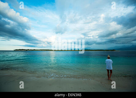 Turista femminile permanente sulla spiaggia al tramonto, Gili Trawangan Isola, guardando verso Gili Meno, isole Gili, Indonesia Foto Stock