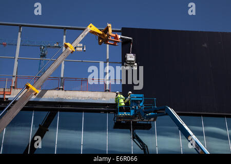 Dockland, Liverpool Waterfront, Merseyside, costruzione in corso sulla facciata del nuovo Kings Dock al centro esposizioni. Un rivestimento esterno essendo fissato utilizzando "Clad Boy' il sollevatore a vuoto dispositivo collegato (fissato con nastro adesivo) a JCB gru 540 170 con due operai, che fissa il rivestimento unità, utilizzando 'Easi-Lifts' estensione benna. Liverpool nel lavoro è sostenere il costruttore principale contraente per lo sviluppo, ISG costruzione, con accesso alla manodopera locale e subappaltatore base, nonché supporto nella compilazione di offerte di lavoro e di apprendistato. Foto Stock