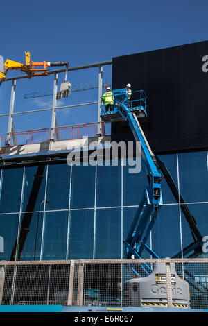 Dockland, Liverpool Waterfront, Merseyside, costruzione in corso sulla facciata del nuovo Kings Dock al centro esposizioni. Un rivestimento esterno essendo fissato utilizzando "Clad Boy' il sollevatore a vuoto dispositivo collegato (fissato con nastro adesivo) a JCB gru 540 170 con due operai, che fissa il rivestimento unità, utilizzando 'Easi-Lifts' estensione benna. Liverpool nel lavoro è sostenere il costruttore principale contraente per lo sviluppo, ISG costruzione, con accesso alla manodopera locale e subappaltatore base, nonché supporto nella compilazione di offerte di lavoro e di apprendistato. Foto Stock