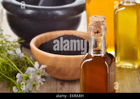 Close-up di nigella sativa olio in una bottiglia, contro di nigella semi e fiori su sfondo di legno. Cumino Nero alle erbe curative. Foto Stock