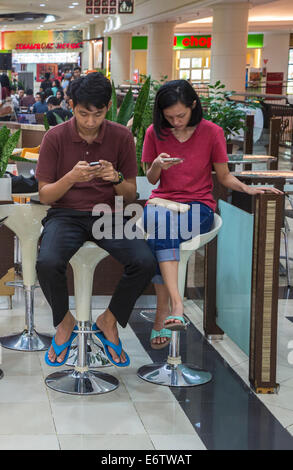 Yogyakarta, Java, Indonesia. Ambarrukmo Shopping Mall. Giovane coppia giavanese controllo telefoni cellulari nel Food Court. Foto Stock