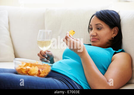 Donna sovrappeso a casa a mangiare patatine e bere il vino Foto Stock