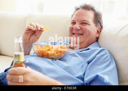 Uomo sovrappeso a casa a mangiare patatine e bere birra Foto Stock