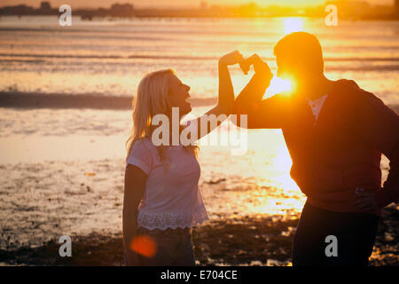 Giovane formando forma di cuore con bracci sulla spiaggia al tramonto Foto Stock