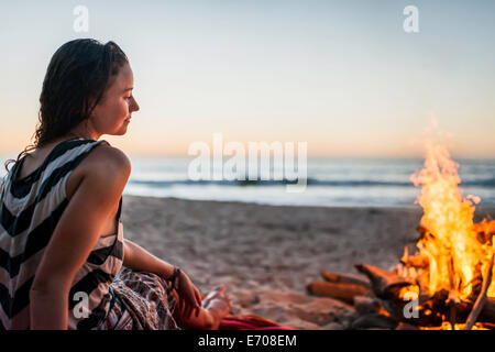Giovane donna seduta al falò sulla spiaggia Foto Stock