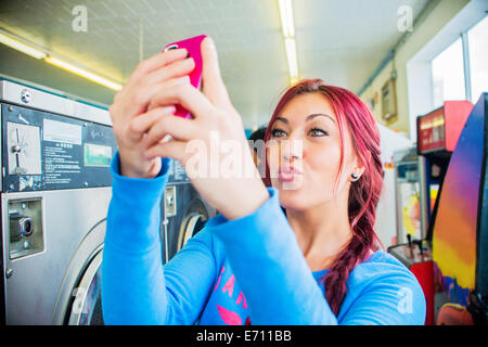 Giovane donna in lavanderia a gettoni, tenendo autoritratto con lo smartphone Foto Stock
