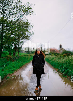 Una donna che cammina su una strada bagnata, indossando un cappotto e cappello. Foto Stock