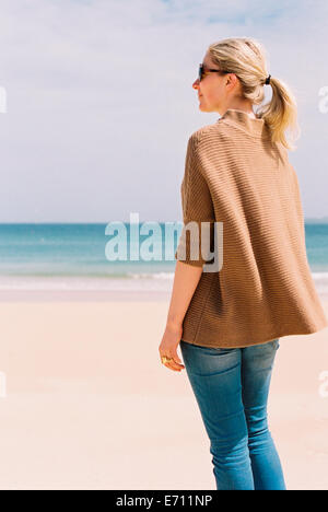 Una donna in una camicia marrone permanente sulla spiaggia che guarda al mare. Foto Stock