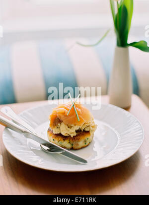 Una piastra con salmone affumicato su un muffin, su un tavolo. Un vaso con fiori e un divano a strisce cuscino. Foto Stock