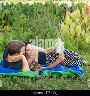 Un giovane uomo e donna seduta su un picnic rug la lettura di un libro con il loro cane piccolo tra loro. Foto Stock