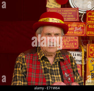 Uomo che indossa vivacemente colorate di rosso, giallo e nero e vestiti con Red Hat vendita segni umoristico alla fiera Foto Stock