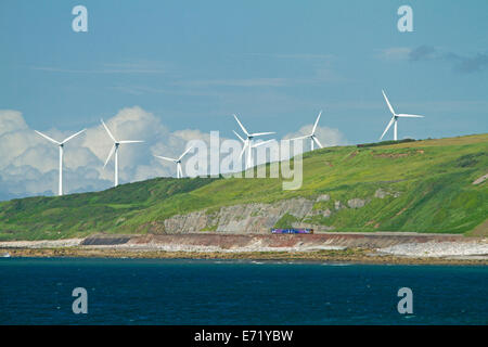 Fila di gigantesche turbine a vento sulla collina verde sulla costa inglese a Whitehaven, Cumbria, contro il cielo blu e rimpicciolendo il treno dei pendolari sulle vie di seguito. Foto Stock