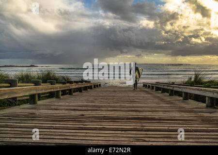 Surfer a piedi lungo una passerella verso la spiaggia con un drammatico sky Foto Stock