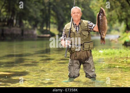 Orgoglioso pescatore in piedi in un fiume e tenendo un pesce e una canna da pesca Foto Stock