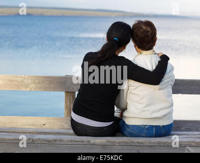 Primo piano della madre con la figlia matura mentre tiene il suo mentre è seduto sulla panca di legno guardando verso l'esterno al lago Foto Stock