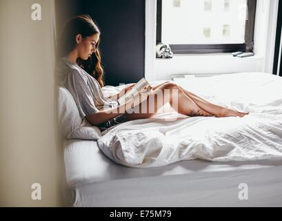 Vista laterale della giovane donna seduta sul suo letto a leggere un libro a casa in camera da letto Foto Stock