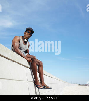 Rilassata giovane uomo seduto su una passeggiata contro il cielo. African maschio modello indossando occhiali da sole e cappello guardando la fotocamera. Foto Stock