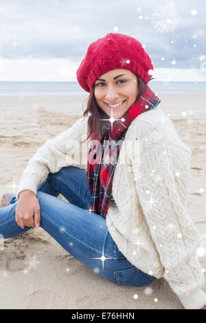 Immagine composita della donna sorridente in eleganti abiti caldi seduto sulla spiaggia Foto Stock
