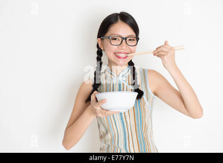 Ritratto di asiatici femminile cinese di mangiare, utilizzando bastoncini holding ciotola di riso, rétro in stile revival cheongsam permanente sulla pianura Foto Stock