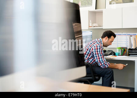 Un uomo che lavora in un ufficio, seduti controllando il suo smart phone. Foto Stock