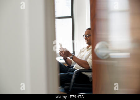 Un uomo seduto in un ufficio, controllando il suo smart phone. Foto Stock