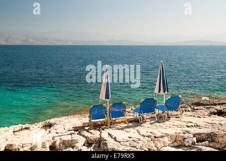 Lettini e ombrelloni (ombrelloni) su una spiaggia rocciosa nell isola di Corfù, Mar Ionio, Grecia Foto Stock