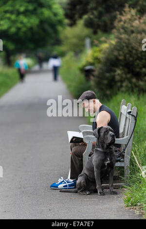 Uomo seduto su una panchina e la lettura di un libro con un cane nel cimitero di Brompton, West Brompton, London, England, Regno Unito Foto Stock