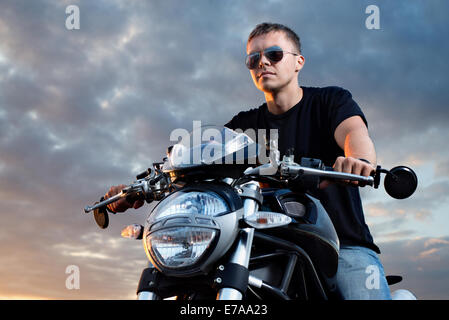 Ritratto romantico bello biker uomo in occhiali da sole si siede su una bicicletta su un Cielo di tramonto sullo sfondo. Copia spazio a sinistra Foto Stock
