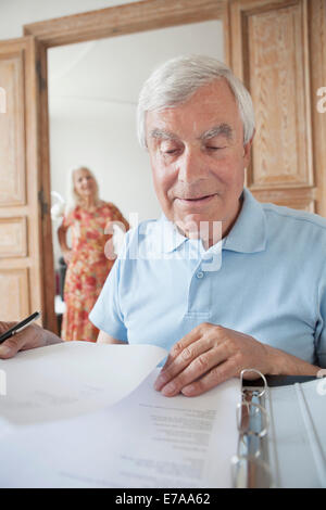 Senior uomo esaminando i documenti finanziari a casa con la donna in background Foto Stock