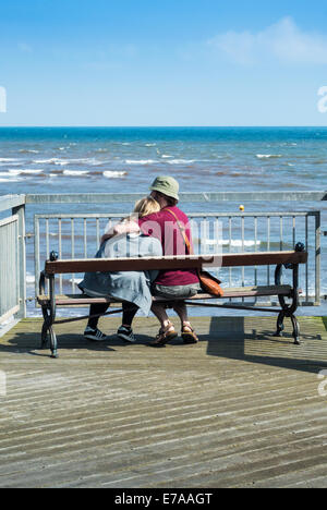 Una giovane coppia sit cuddling reciprocamente in Teignmouth Pier a guardare il mare. Foto Stock