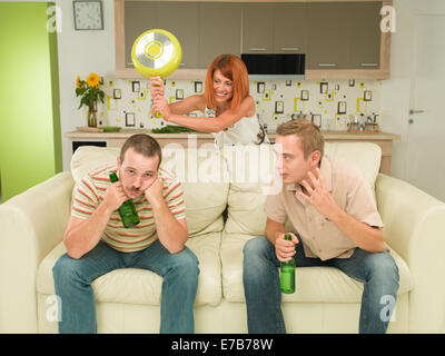 Due uomini caucasici seduta sul lettino tenendo le bottiglie di birra, avente la conversazione, donna in background cercando di colpire uno di loro con Foto Stock