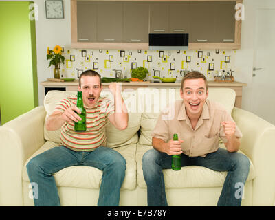 Due uomini eccitati seduti sul divano guardando la televisione, in cucina, azienda orsi e urlando Foto Stock