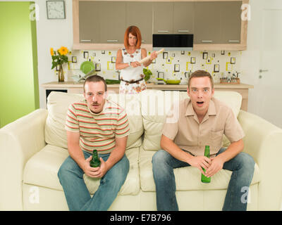 Due uomini seduti sul divano guardando la televisione, in cucina, tenendo le birre e deliberando sorpreso con un arrabbiato donna in background Foto Stock