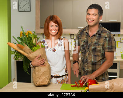 Vista frontale della coppia felice in cucina, donna holding sacchetto di carta con negozi di generi alimentari in piedi accanto a un uomo tritare il peperone rosso e smi Foto Stock