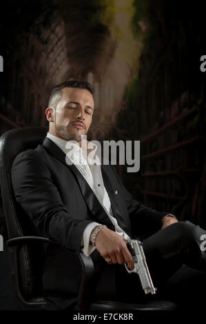 Elegante uomo seduto in una sedia tenendo la pistola su sfondo scuro Foto Stock