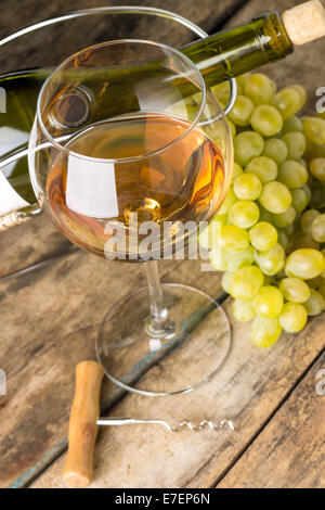 Wineglass con vino bianco, bottiglia, cavatappi e il grappolo intorno su uno sfondo di legno Foto Stock