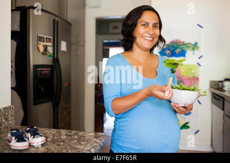 Incinta donna ispanica mangiare insalata in cucina Foto Stock