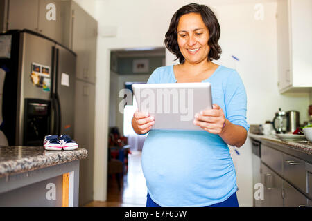 Incinta donna ispanica utilizzando computer tablet in cucina Foto Stock