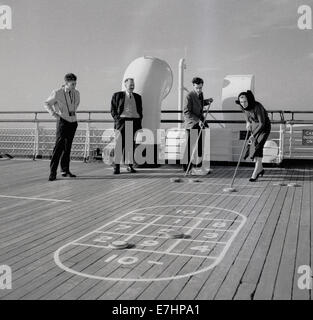 1950s. Un quadro storico di tre maschi e una femmina di passeggeri su una nave da crociera godendo un mazzo di gioco shove il puck. Foto Stock