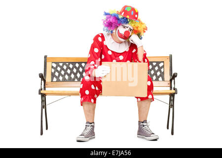 Triste clown maschile seduto su una panca in legno e in possesso di un cartone vuoto segno isolato su sfondo bianco Foto Stock