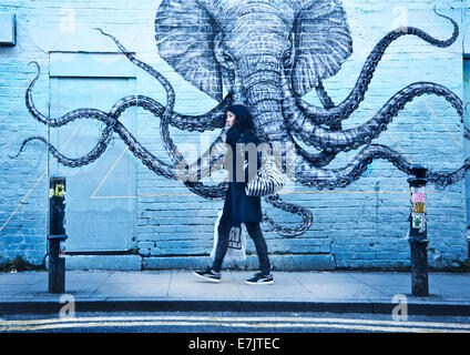 Una ragazza camminare sulla strada che passa un graffito con un elefante/octopus su un muro di mattoni. Foto Stock