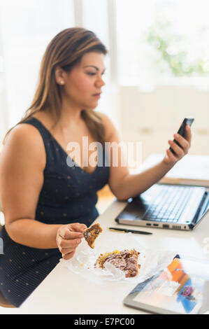 Donna di mangiare croissant e utilizzando il telefono cellulare in ufficio Foto Stock