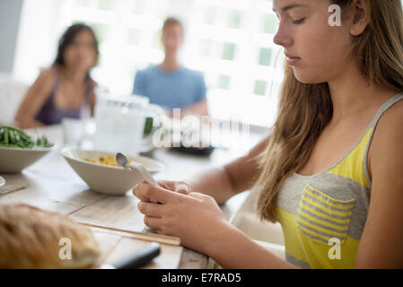 Una giovane ragazza seduti controllando il suo smart phone a un tavolo da pranzo. Due persone in background. Foto Stock